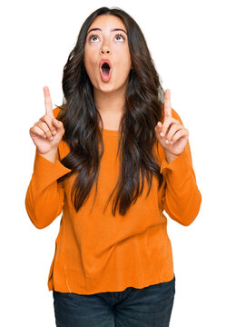 Beautiful brunette young woman wearing casual orange sweater amazed and surprised looking up and pointing with fingers and raised arms.