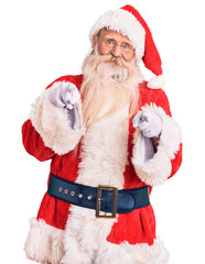 Old senior man with grey hair and long beard wearing traditional santa claus costume pointing fingers to camera with happy and funny face. good energy and vibes.