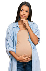 Beautiful hispanic woman expecting a baby showing pregnant belly serious face thinking about...