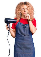 Young blonde woman with curly hair wearing hairdresser apron and holding dryer blow serious face thinking about question with hand on chin, thoughtful about confusing idea
