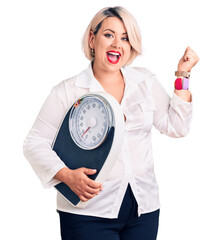 Young blonde plus size woman holding weighing machine screaming proud, celebrating victory and...