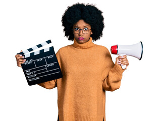 Young african american woman holding video film clapboard and megaphone skeptic and nervous,...