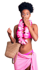 Young african american woman wearing bikini and hawaiian lei covering mouth with hand, shocked and...