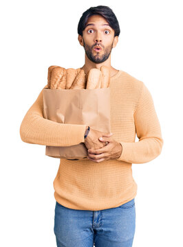 Handsome hispanic man holding paper bag with bread scared and amazed with open mouth for surprise, disbelief face