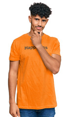 Fototapeta premium Young arab man wearing tshirt with happiness word message looking confident at the camera smiling with crossed arms and hand raised on chin. thinking positive.