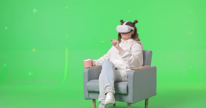 Shot on green screen background. VR or AR online at home. Cute girl in virtual reality headset in chair. Woman clicks invisible buttons pulls virtual sliders and drinking from a cup. Internet fun.