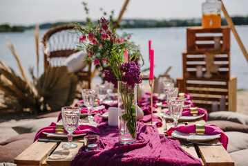 Picnic on beach at sunset in rustic style, boho, food, wine conception. Decor with blanket, pillows, wooden decor, candles, pink flowers, dry leaves, straw chair. Bachelorette party, birthday, wedding