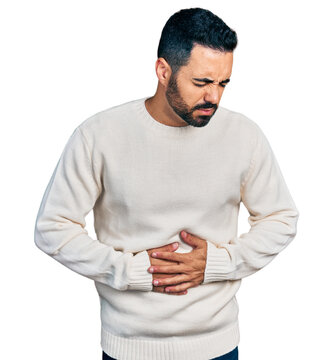 Young hispanic man with beard wearing casual white sweater with hand on stomach because indigestion, painful illness feeling unwell. ache concept.