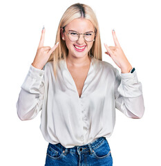 Beautiful blonde woman wearing elegant shirt and glasses shouting with crazy expression doing rock symbol with hands up. music star. heavy music concept.