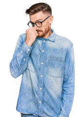 Young redhead man wearing casual denim shirt smelling something stinky and disgusting, intolerable smell, holding breath with fingers on nose. bad smell