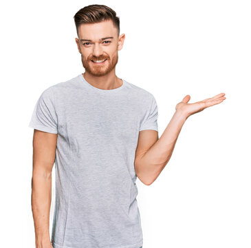 Young redhead man wearing casual grey t shirt smiling cheerful presenting and pointing with palm of hand looking at the camera.