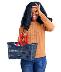Beautiful african american woman holding supermarket shopping basket smiling happy doing ok sign with hand on eye looking through fingers