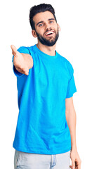Fototapeta premium Young handsome man with beard wearing casual t-shirt smiling cheerful offering palm hand giving assistance and acceptance.