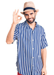 Naklejka premium Young handsome man with beard wearing summer hat and striped shirt smiling positive doing ok sign with hand and fingers. successful expression.