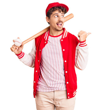 Young handsome man with curly hair playing baseball holding bat and ball pointing thumb up to the side smiling happy with open mouth