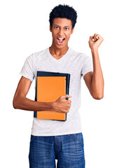 Young african american man holding book screaming proud, celebrating victory and success very excited with raised arms