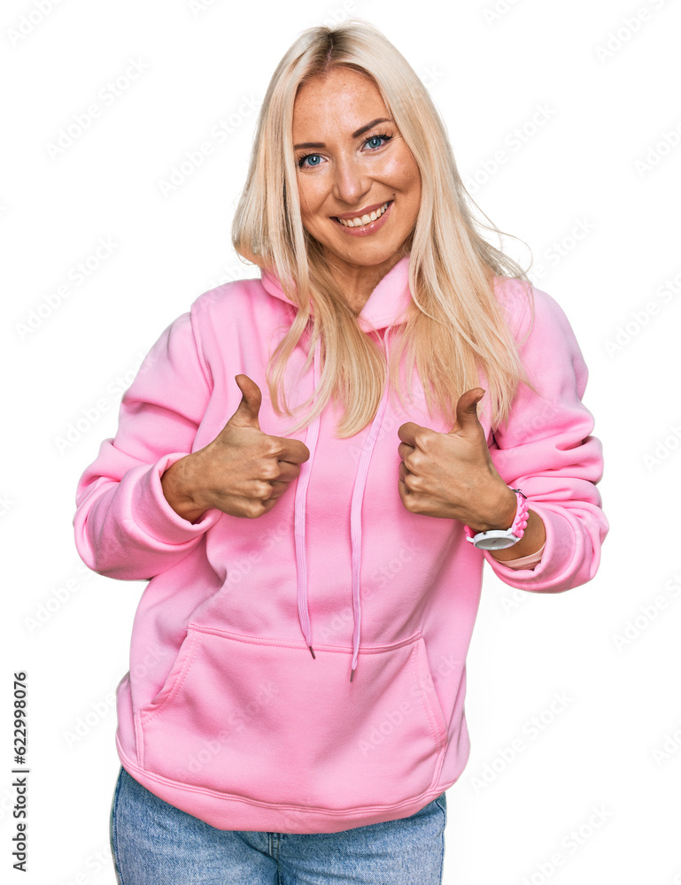 Sticker young blonde woman wearing casual sweatshirt success sign doing positive gesture with hand, thumbs u