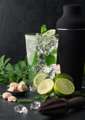 Glass of Mojito cocktail with ice cubes mint and lime on black board with black shaker and wooden squeezer and fresh limes with cane sugar.