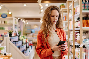 Young customer using smartphone while doing shopping in cosmetics store