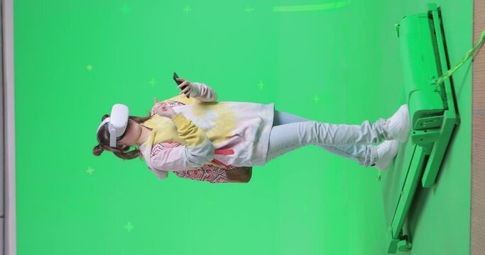 Vertical shot on green screen background. VR or AR online walk. Girl walks in augmented reality headset and uses a mobile smartphone. Walking on a treadmill at 45 degrees to camera.