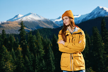 Naklejka premium Woman hiker standing on the mountain looking at nature happiness with a view of snowy mountains and trees in yellow raincoat and cap travel autumn and hiking in the mountains at sunset freedom