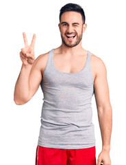 Naklejka premium Young handsome man wearing swimwear and sleeveless t-shirt smiling with happy face winking at the camera doing victory sign with fingers. number two.