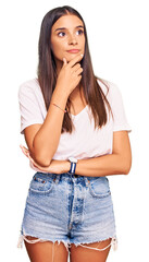 Young hispanic woman wearing casual white tshirt with hand on chin thinking about question, pensive expression. smiling with thoughtful face. doubt concept.