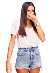 Young hispanic woman wearing casual white tshirt smelling something stinky and disgusting, intolerable smell, holding breath with fingers on nose. bad smell
