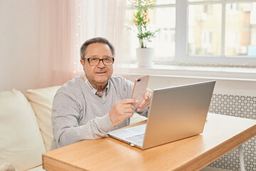 Fototapeta premium Cheerful senior man has phone conversation, chatting on the smartphone working with a laptop remotely from home.
