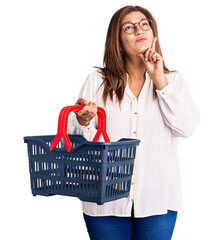 Middle age latin woman holding supermarket shopping basket serious face thinking about question with hand on chin, thoughtful about confusing idea