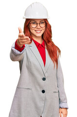 Young redhead woman wearing architect hardhat smiling friendly offering handshake as greeting and welcoming. successful business.