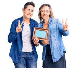 Couple of women holding empty frame smiling looking to the camera showing fingers doing victory sign. number two.