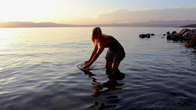 Beautiful young woman turning with hat and splashing water from sea. Enjoying vacation, slow motion in sunset. Croatia, island Krk