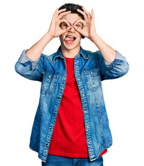 Young caucasian boy with ears dilation wearing casual denim jacket doing ok gesture like binoculars sticking tongue out, eyes looking through fingers. crazy expression.