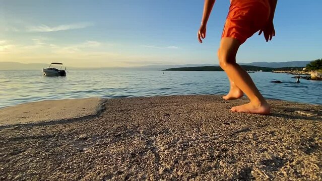 Young caucasian boy in orange shorts jumping into sea with boat. Slow motion, holiday, vacation background. Croatia, island Krk