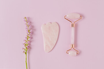Pink quartz roller and gouache scraper with a flower on a purple isolated background. The concept of natural skin care. Massage for nutrition, wrinkling, tightening of contours of the face