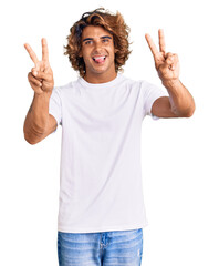 Young hispanic man wearing casual white tshirt smiling with tongue out showing fingers of both hands doing victory sign. number two.