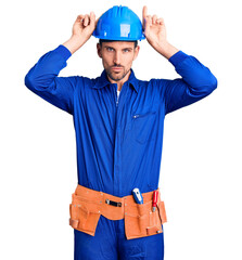 Young handsome man wearing worker uniform and hardhat doing funny gesture with finger over head as bull horns