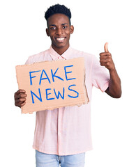 Young african american man holding fake news banner smiling happy and positive, thumb up doing...
