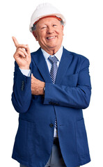 Senior handsome grey-haired man wearing suit and architect hardhat with a big smile on face, pointing with hand and finger to the side looking at the camera.