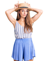 Young beautiful blonde woman wearing summer hat doing funny gesture with finger over head as bull horns