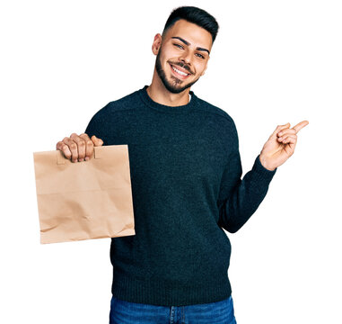 Young hispanic man with beard holding take away paper bag smiling happy pointing with hand and finger to the side