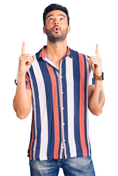 Young handsome hispanic man wearing casual  summer shirt amazed and surprised looking up and pointing with fingers and raised arms.