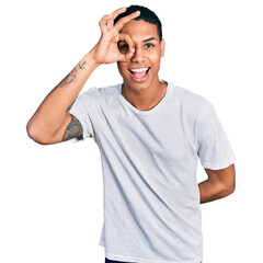 Fototapeta premium Young hispanic man wearing casual white t shirt doing ok gesture with hand smiling, eye looking through fingers with happy face.