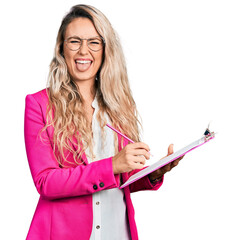 Young blonde woman writing on clipboard sticking tongue out happy with funny expression.