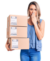 Young beautiful blonde woman holding delivery boxes covering mouth with hand, shocked and afraid...