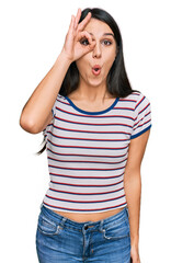 Obraz na płótnie Canvas Young hispanic girl wearing casual striped t shirt doing ok gesture shocked with surprised face, eye looking through fingers. unbelieving expression.