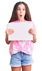 Cute hispanic child girl holding blank empty banner scared and amazed with open mouth for surprise, disbelief face