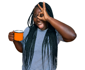 Young black woman with braids drinking a cup coffee smiling happy doing ok sign with hand on eye looking through fingers
