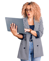 Young blonde woman with curly hair working using laptop scared and amazed with open mouth for surprise, disbelief face